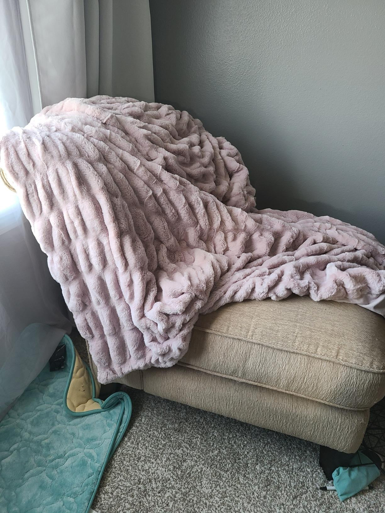A pink textured Aruva SereneWeave blanket draped over a cozy beige armchair next  to a window with sheer curtains in a room with sage-colored walls and a teal rug.