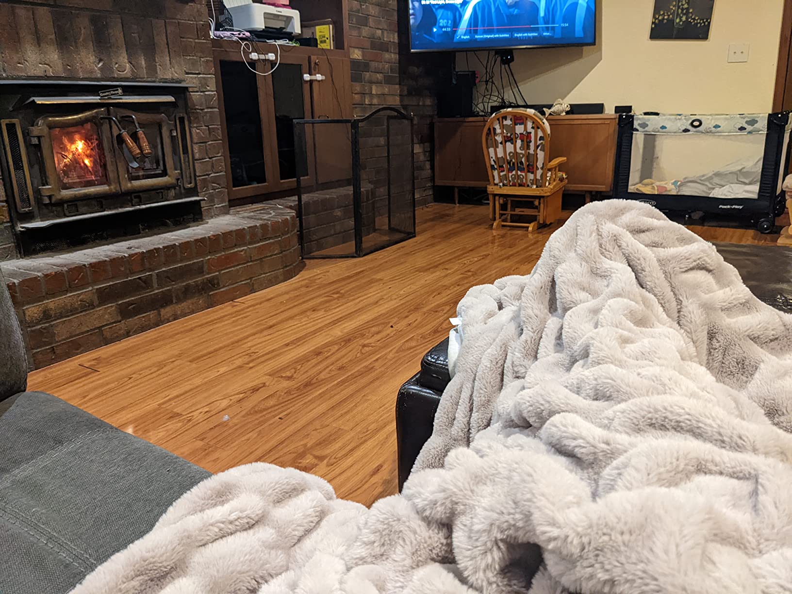 Customer photo showing someone relaxing on their living room sofa with an Aruva SereneWeave throw keeping their legs warm with a blazing fireplace and TV in the background.