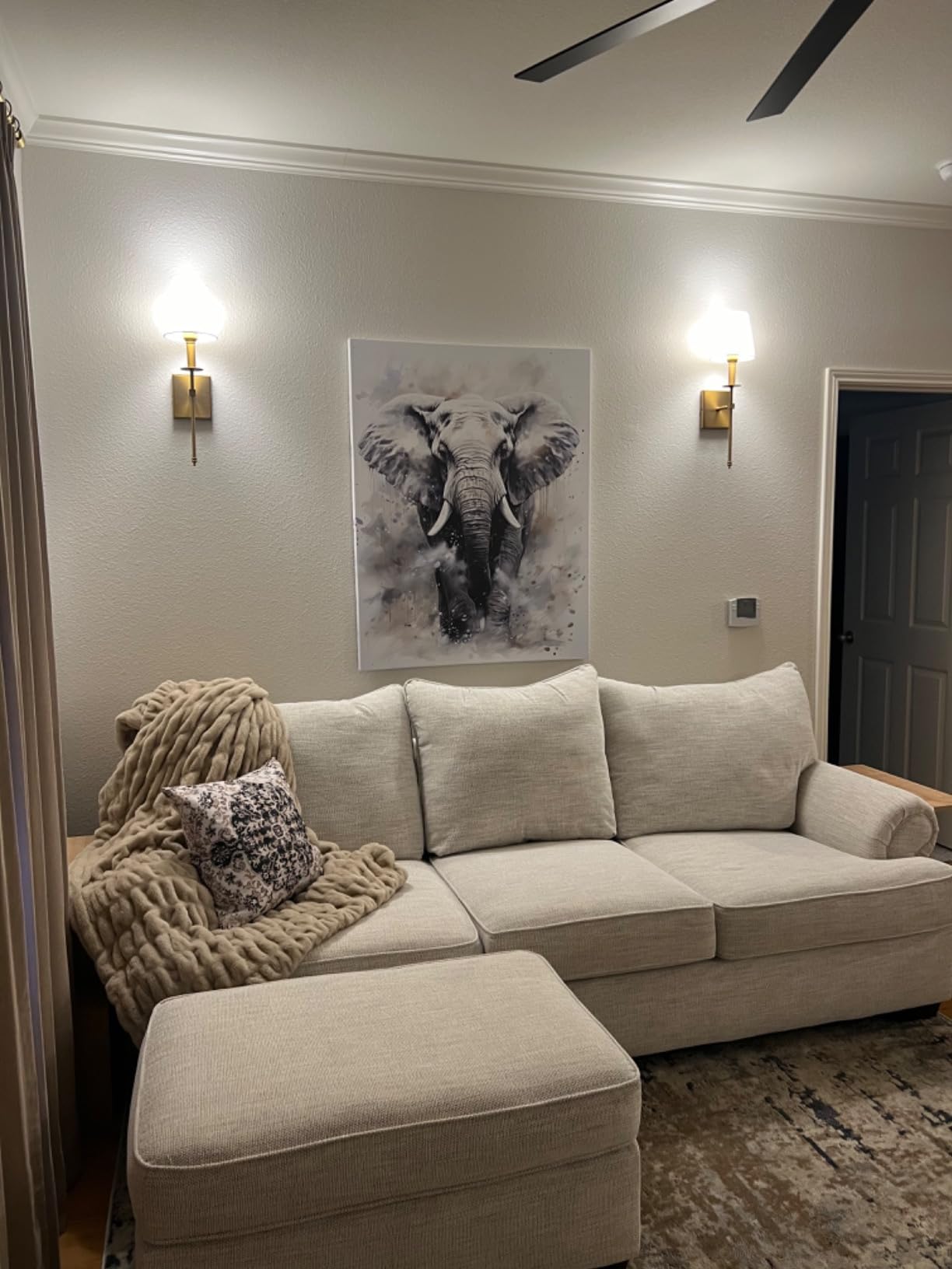 Customer photo showing Aruva SereneWeave throw decorating a Beige sectional sofa with ottoman in a living room setting, featuring wall art of an elephant.